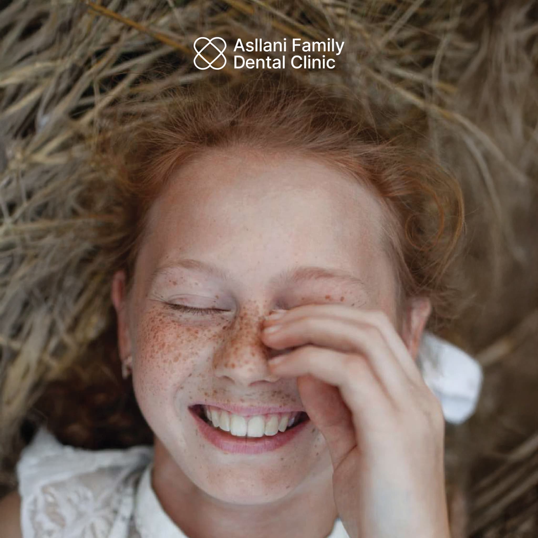 A close-up portrait of a smiling young girl with freckles, with the Asllani Family Dental Clinic logo lockup applied in the upper corner — showing the brand's warmth in a real lifestyle context.
