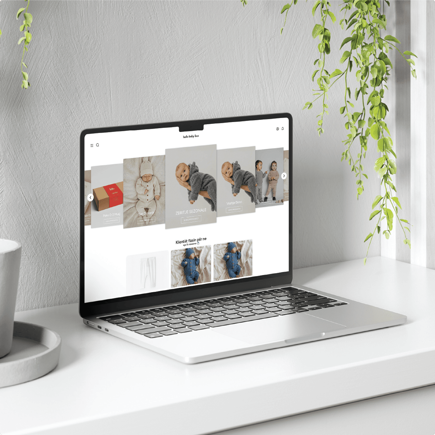 Hello Baby Box homepage on a laptop in a soft natural-lit setting with hanging greenery — showing product imagery and customer testimonials.