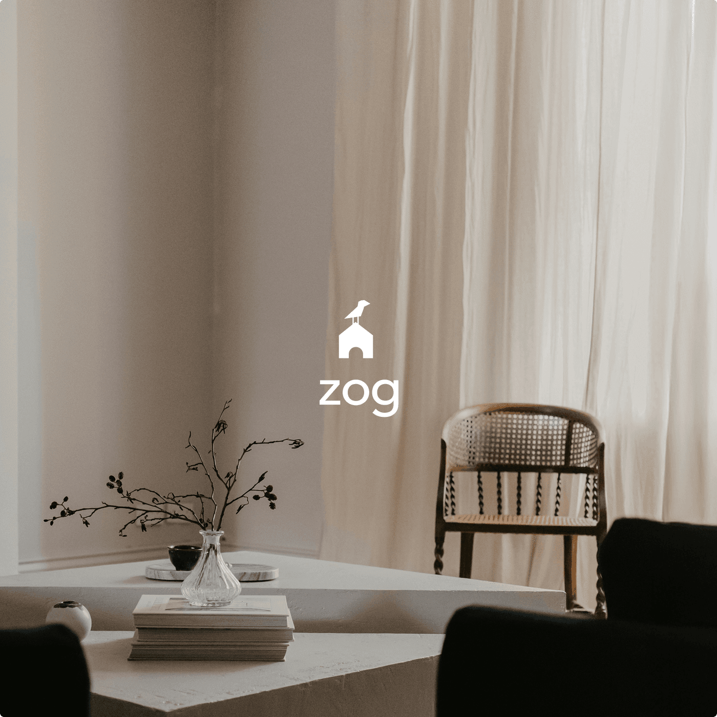 Ein ruhiges Interieur — beige Wände, leichte Vorhänge, ein Korbstuhl aus Holz, ein Couchtisch aus Stein mit Büchern und einer Vase mit kahlen Zweigen — mit der ZOG-Wortmarke und dem Vogel-auf-Bogen-Monogramm in Weiß im oberen Bildbereich.